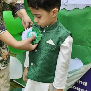 A teacher pins a handmade green and white rosette to a young student's waistcoat during Independence Day celebrations
